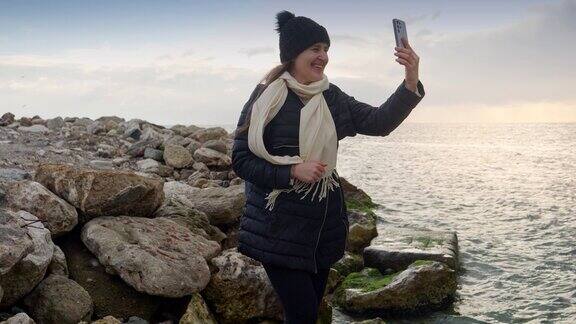 戴着帽子和围巾的微笑女子站在海边的悬崖上用智能手机进行视频通话旅游、旅行、旅行、假期和冬季度假