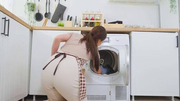 亚洲美女把脏衣服放在家里的洗衣机里漂亮的女孩穿着围裙坐在地板上高兴地把衣服放进家里的洗衣机里洗居住房屋保持概念