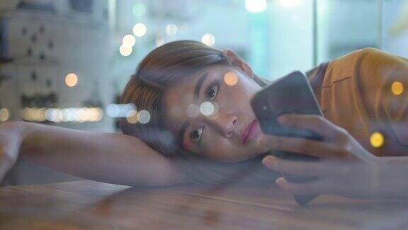 4K孤独年轻美丽的亚洲女人坐在窗前躺在桌子上晚上在咖啡馆用智能手机上网悲伤的少女望着窗外灯火通明的城市街道