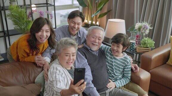 笑容满面的亚洲大家庭和孩子们坐在客厅的沙发上一起在手机上自拍视频快乐的父母和孩子们在家里用智能手机进行有趣的视频通话