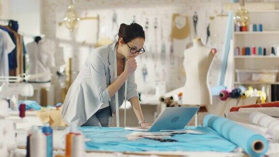 美丽的女设计师使用笔记本电脑在她的桌子上覆盖着各种织物和缝纫项目她的工作室是Sunny有人体模型挂衣服和草图钉在墙上