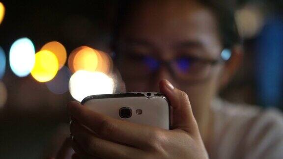 戴眼镜的亚洲女性用智能手机在城市的夜晚灯光背景下发短信