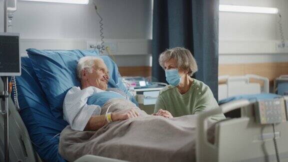 医院病房:躺在床上的老人戴着口罩的美丽妻子探望心爱的丈夫支持他坐在旁边手牵着手幸福的夫妻交谈手术后恢复