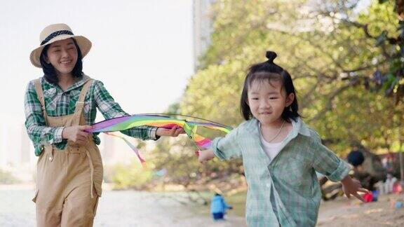 小亚洲儿子在沙滩上玩五彩缤纷的风筝美丽的亚洲母亲拿着风筝跟着儿子跑在风筝随风飘动前支撑着他们很开心有微笑的脸与家人共度美好时光