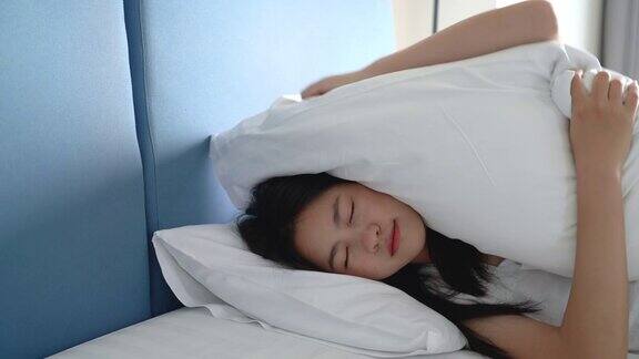 亚洲女人失眠试图用盖住耳朵的枕头睡觉她听到失眠和烦躁的噪音问题在酒店房间的床上醒来