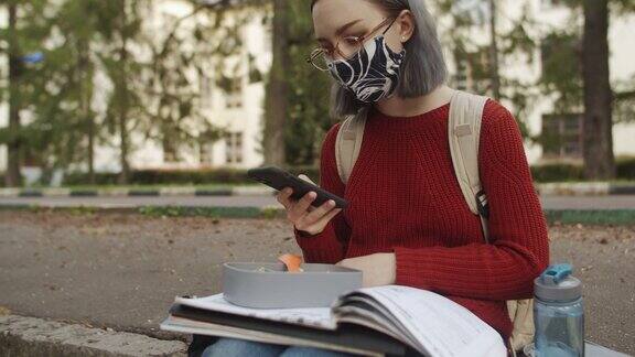 在大学校园里年轻的学生戴着智能手机戴着可重复使用的防护口罩在学校附近行走