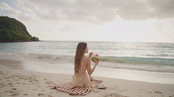 亚洲年轻女子穿着泳衣喝着椰子汁坐在沙滩上看书