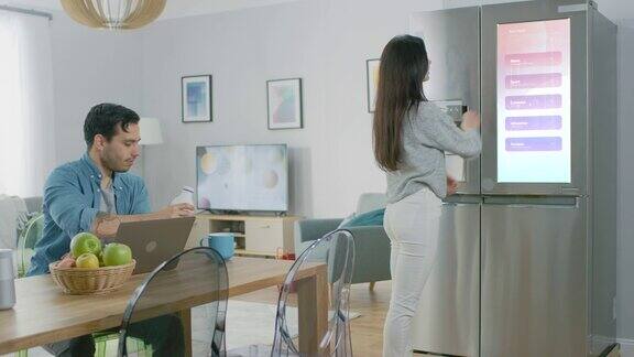 美丽的年轻女子打开冰箱给了她的男朋友一个奶瓶然后她检查智能冰箱门上的未来数字待办事项清单
