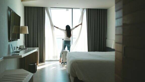 肖像的女游客站在近窗口看美丽的景色与她的行李在酒店卧室登记后