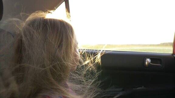 年轻的白人女孩望着车窗外的地平线打开车窗强风吹动她的头发近距离手持式拍摄乘汽车旅行秋天日落背光太阳耀斑