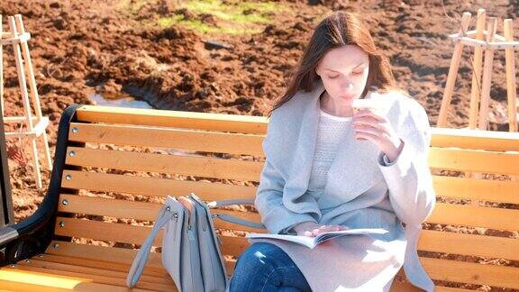 年轻漂亮的黑发女学生坐在春天公园的长椅上一边看书一边喝咖啡