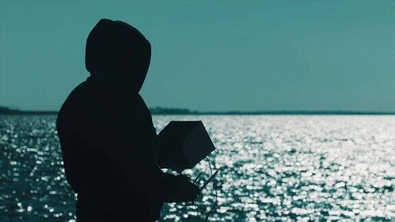 在湖岸戴着兜帽的男子使用无人机控制器的剪影