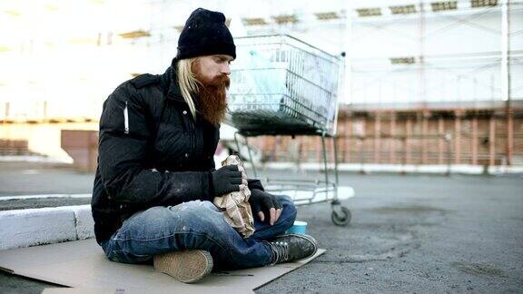 在寒冷的冬天一个无家可归的年轻人穿着脏衣服坐在街道上的购物车旁喝酒