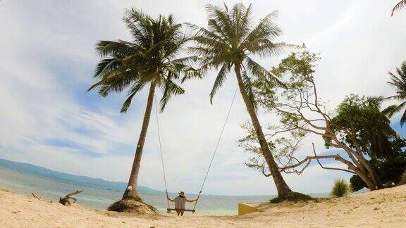 快乐的年轻女子在椰子树上荡秋千看着令人惊叹的热带海滩