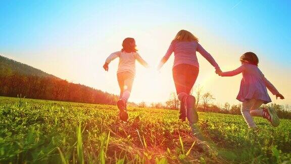 SLOMOTS女人和她的两个女儿手牵着手在阳光明媚的草地上奔跑