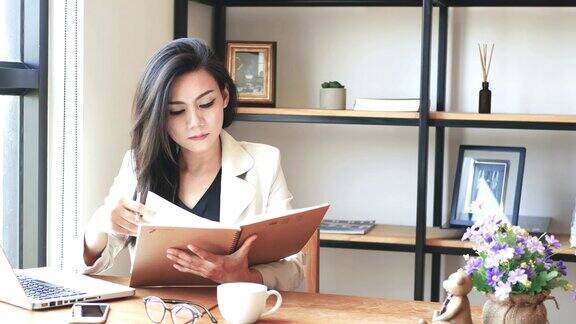 4K视频美丽的亚洲商务女性坐在电脑笔记本电脑阅读和翻页笔记本和微笑在咖啡馆咖啡厅在城市的早晨商务人士的生活方式