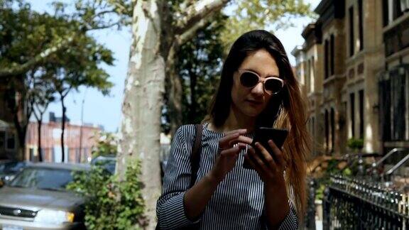 年轻漂亮的女人在阳光明媚的夏天戴着太阳镜走在外面使用智能手机的肖像慢动作
