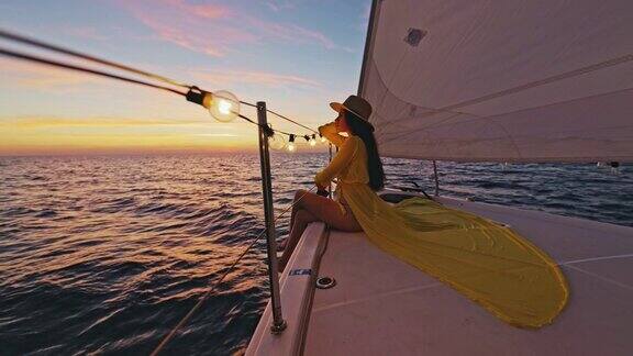 一个穿着黄色长裙的女人坐在夕阳下航行的帆船的甲板上