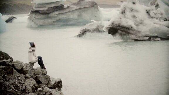 年轻迷人的女人坐在冰泻湖的岩石上游客在lopapeysa探索冰岛的风景