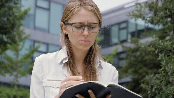 年轻的女学生戴着眼镜坐在户外的长椅上写笔记