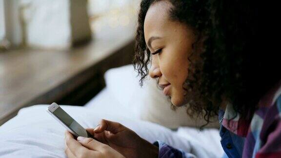 非洲女孩躺在家里的床上用智能手机看网上照片的特写