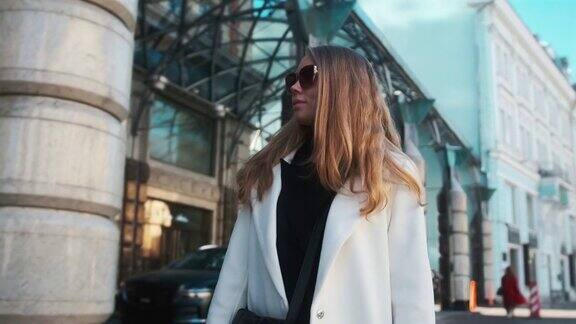 一个戴着眼镜穿着白色外套的女孩走过城市的商业区在豪华轿车和公寓的背景下