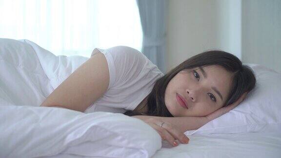 美丽的亚洲女人肖像与迷人的微笑享受新鲜柔软的床上用品亚麻布床垫在白色的卧室现代公寓可爱的亚洲女孩睡觉休息晚安睡眠