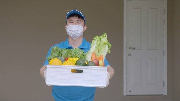 在居家隔离期间一名亚洲快递员戴着口罩和手套拿着装满食品、水果、蔬菜和饮料的杂货箱站在客房前的货车旁