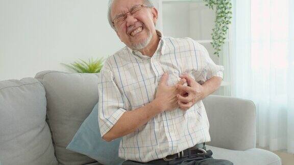 资深男性亚洲人遭受严重的疼痛在他的胸部心脏病发作坐在家里的沙发上老人把他的手放在他的左胸口因为他的心突然疼痛心脏病