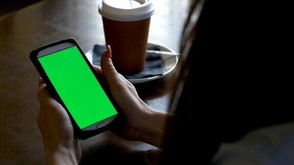 一个女人的手坐在桌子上拿着绿色屏幕的智能手机喝着咖啡