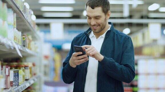 在超市:英俊的男人使用智能手机站在罐头食品区微笑