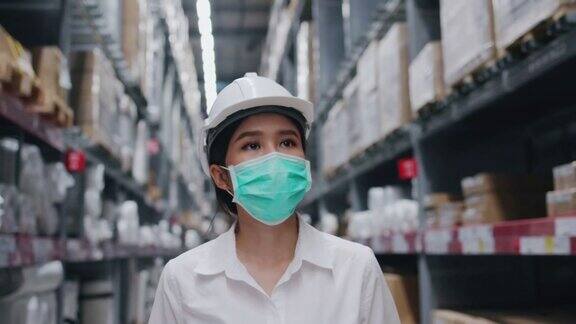 亚洲女仓库工人戴安全帽面罩正在工厂检查产品女商人在剪贴板上储存货架信息在工厂里戴口罩预防新冠肺炎和灰尘
