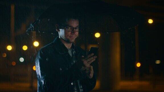 一名戴眼镜、穿着牛仔裤外套和方衬衫的年轻白人男子在雨伞下使用智能手机外面很黑还在下雨