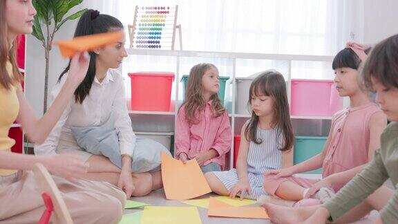 孩子们坐在地板上折纸旁边是他们的幼儿园老师老师一边和孩子们说话一边也在女孩的纸上写东西