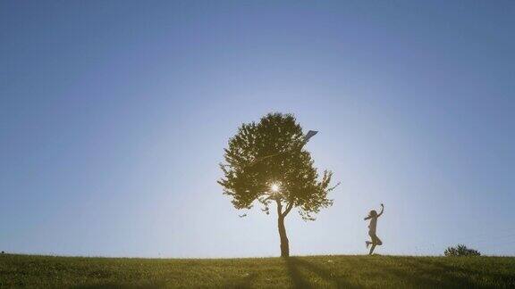 拿着风筝的女孩剪影慢动作拍摄在草地上与美丽的树在蓝色的背景