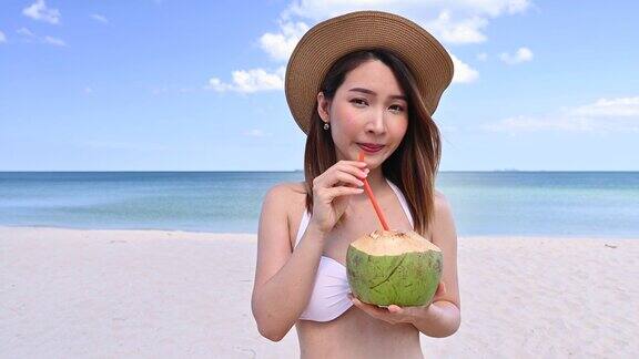 身穿比基尼的亚洲女子在热带海滩享受新鲜的椰子汁