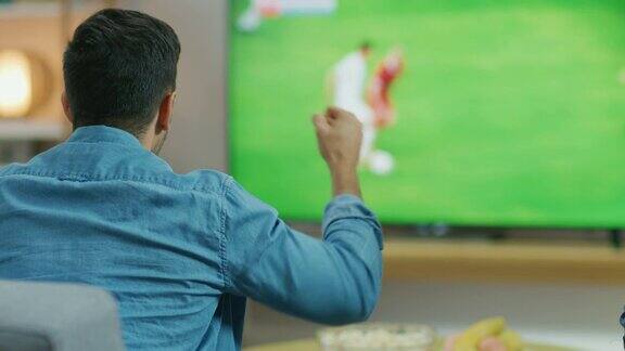 在家乡体育迷在电视上观看重要的足球比赛他积极地挥拳为他的球队欢呼舒适的客厅桌上放着零食和饮料