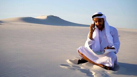 在炎热的日子里一个自信的阿拉伯阿拉伯建筑工人坐在沙地上用手机与客户交谈