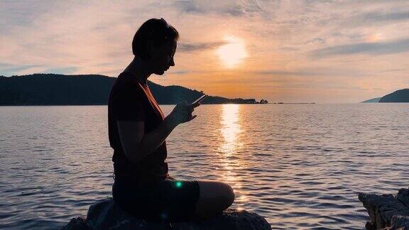 年轻浪漫的女人坐在海边的岩石上欣赏壮观的海景和火红的日落她拿出手机拍下了美丽的风景
