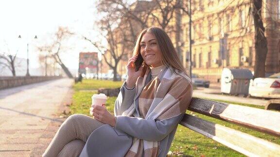 微笑的年轻女子喝着外卖咖啡在城市公园里用智能手机聊天