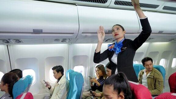 美丽的亚洲空乘人员穿着制服帮助乘客将背包和行李放入头顶的行李架并关闭而乘客在客舱里坐着使用智能手机并准备起飞