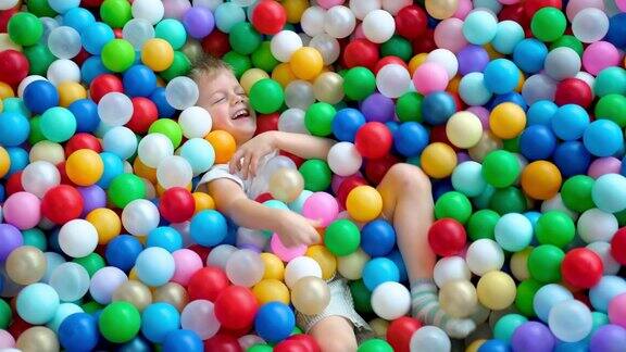 金发小男孩把球扔向不同的方向躺在游戏中心的一个大的干燥的划水池里的彩色塑料球上微笑在相机在游戏室玩得开心休闲活动