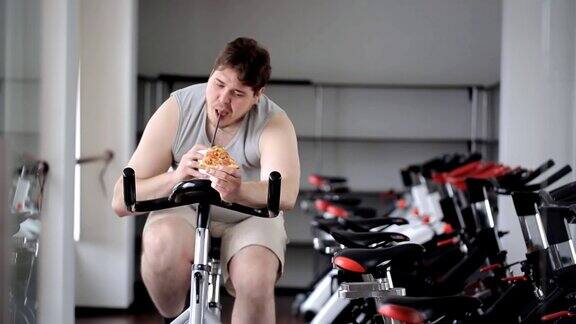 胖子转动着健身自行车的踏板手里拿着一杯饮料和一片披萨