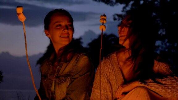 两个年轻的白种女孩晚上坐在大自然的火旁煮着棉花糖微笑着吃着