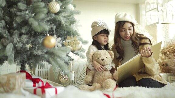 可爱的小女孩和妈妈在客厅的圣诞树旁读童话书