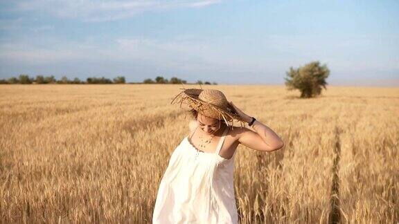 穿着白衣服的漂亮年轻女子在麦田里奔跑微笑着快乐的女性跑着拿着她的草帽在她的头上前视图