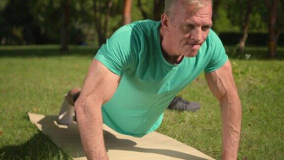 一个穿着蓝绿色t恤的金发体育教练在瑜伽垫上支撑着平板我们从正面看到他