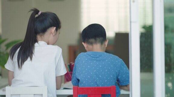 后视图:在学校因新冠肺炎疫情而关闭期间亚洲兄弟姐妹在家里客厅的桌子上肩并肩做作业