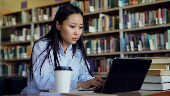 年轻的亚洲学生女孩坐在大学图书馆的一堆书的桌子上在笔记本电脑上工作喝咖啡微笑着看着别处准备考试