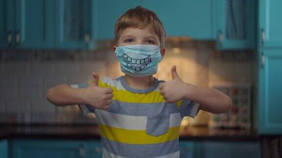 照片中的男孩戴着医用面具微笑着看着相机竖起大拇指孩子戴着有趣的医用口罩在家自我隔离病毒停止流行